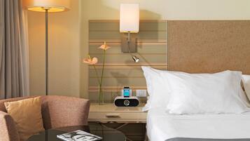 Una habitación de hotel con cama, mesita de noche, lámpara, un jarrón con flores, una mesa con revista y un radio incorporado a la pared.