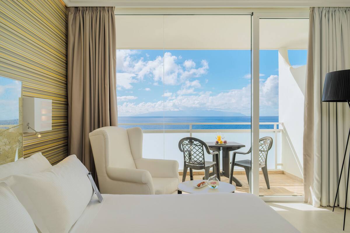 Habitación de hotel con cama, una silla, una mesa con fruta y un balcón con vista al mar.