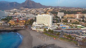 Un hotel frente al mar con varios pisos, una zona de playa con sombrillas y vistas a la ciudad y montañas circundantes.