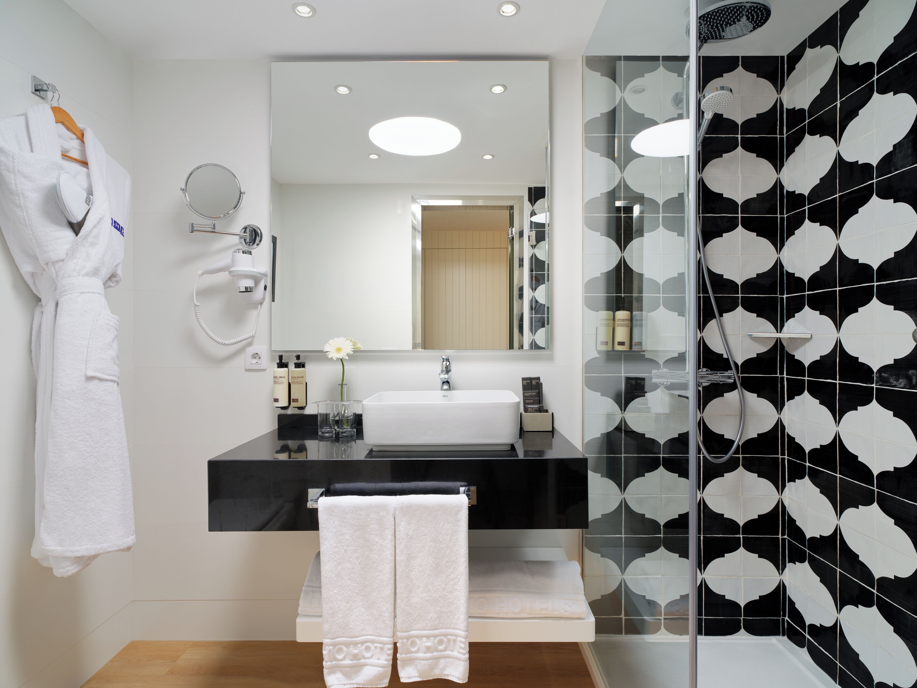 A modern bathroom with a black and white tiled shower, a white sink, and a robe hanging on the wall.