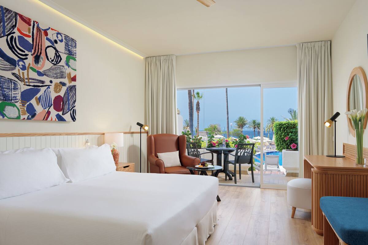 Habitación de hotel con una cama grande, un escritorio, una silla, una mesita y vistas a la piscina y palmeras.