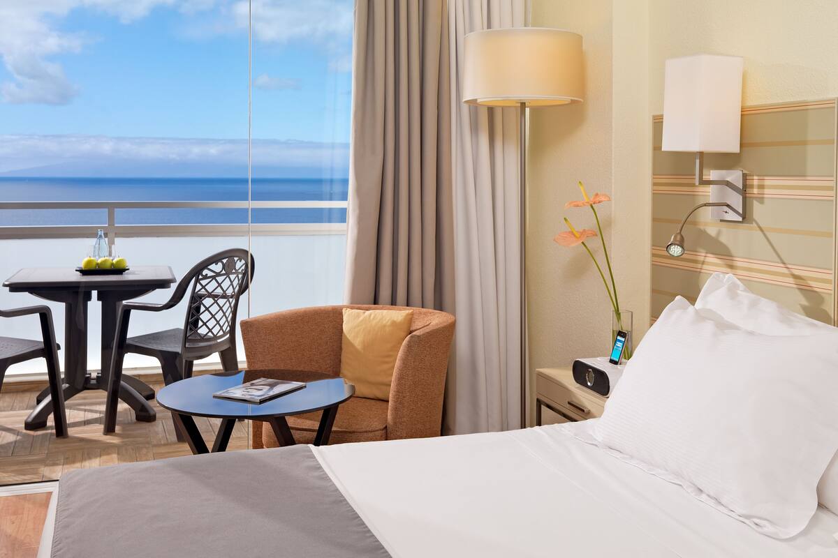 Habitación de hotel con cama, escritorio, silla, mesita, lámpara y vista al océano.