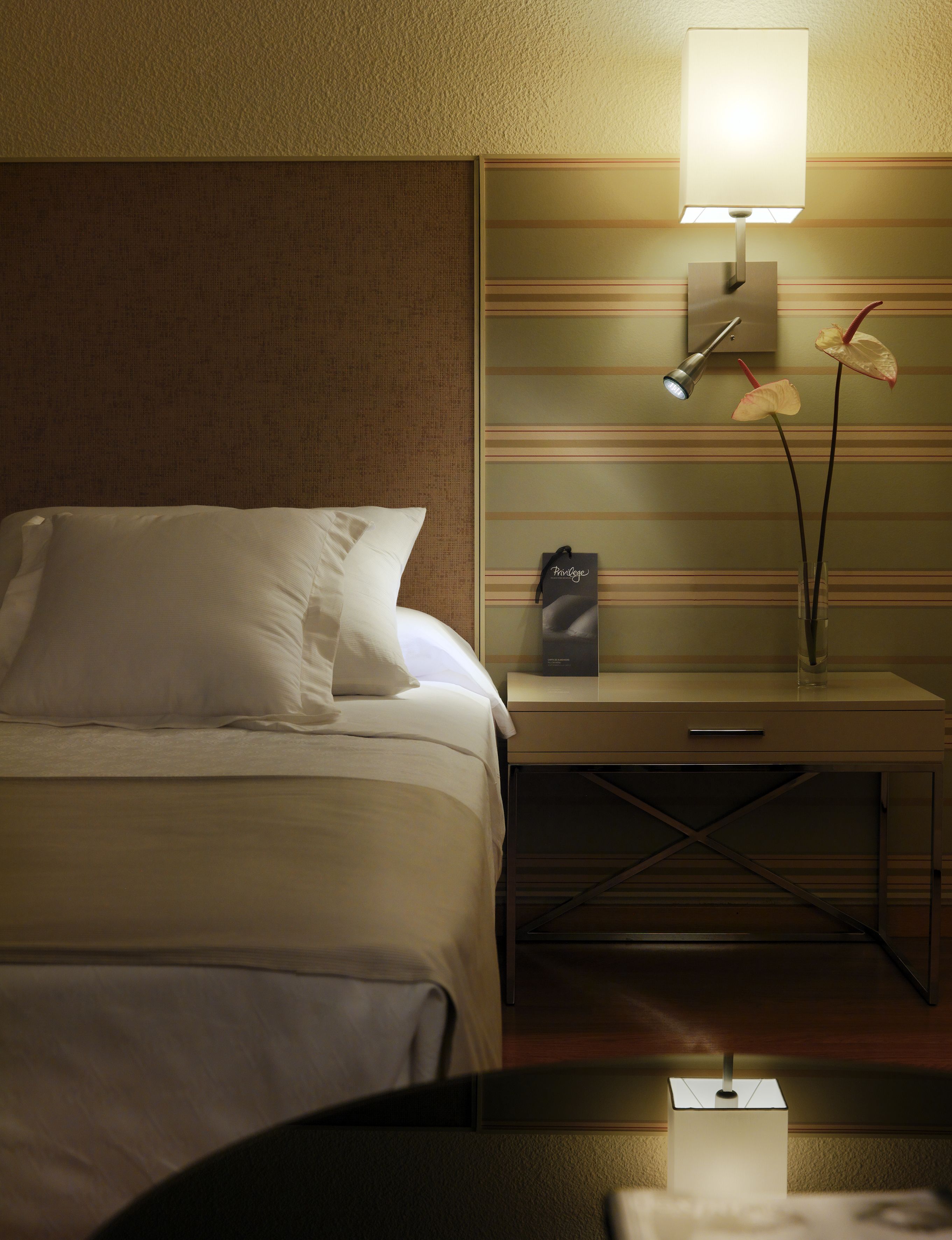 Una habitación de hotel con cama, mesita de noche, lámpara y un florero con flores.
