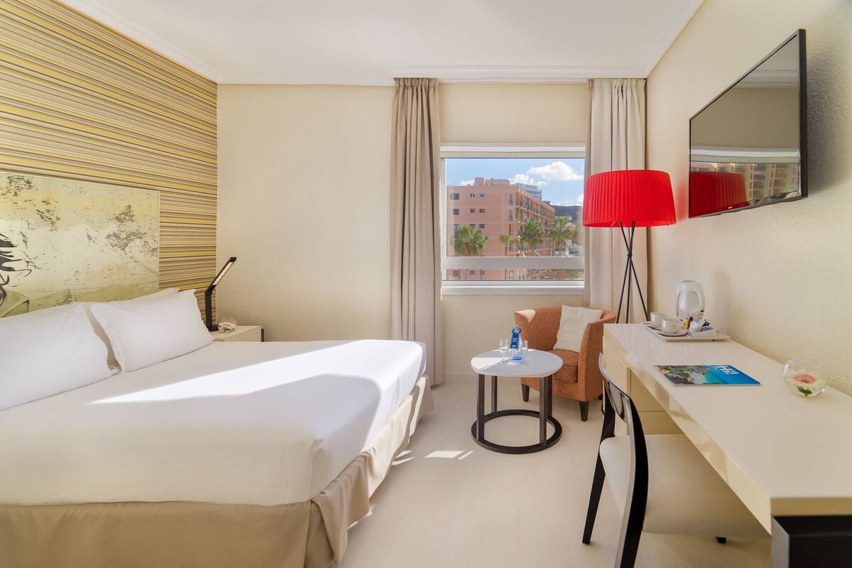 Habitación de hotel con cama, escritorio, silla, mesita, lámpara y ventana con vista a edificios.