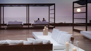 Un espacio al aire libre tranquilo con terraza de madera, sofás blancos acolchados y velas.