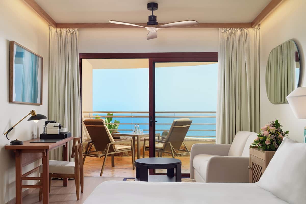 A hotel room with a bed, a desk with a lamp, a chair, a small table, and a view of the ocean.