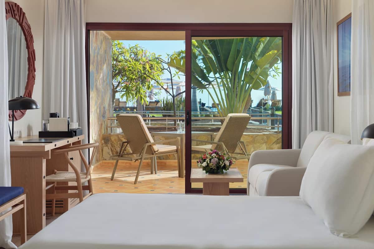 A hotel room with a bed, a desk with a lamp, two chairs, a small table with a flower vase, and a sliding glass door leading to an outdoor area with chairs and a view of greenery.