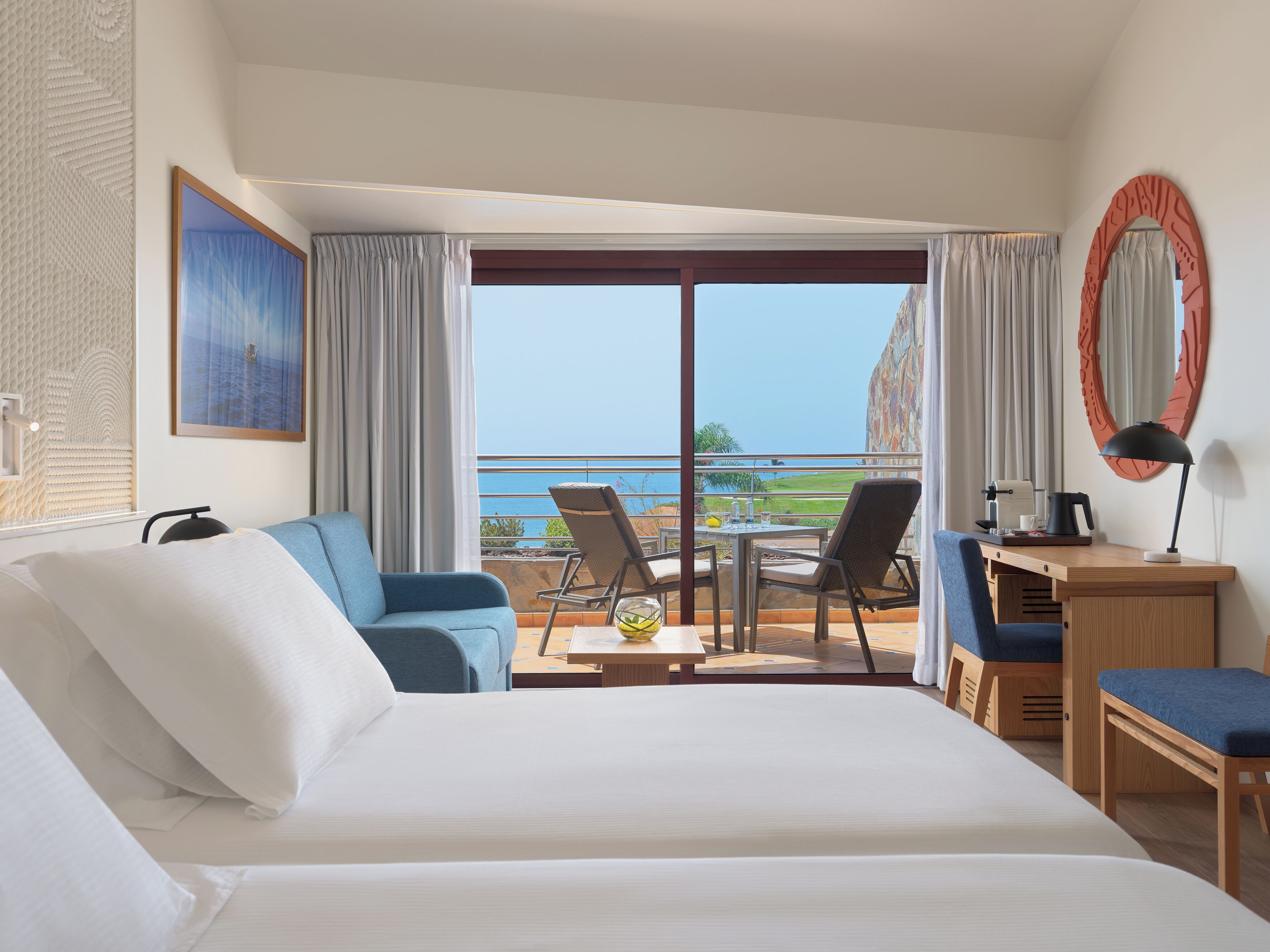 Habitación de hotel con una cama grande, un escritorio con silla, una mesita con lámpara y vistas al océano.
