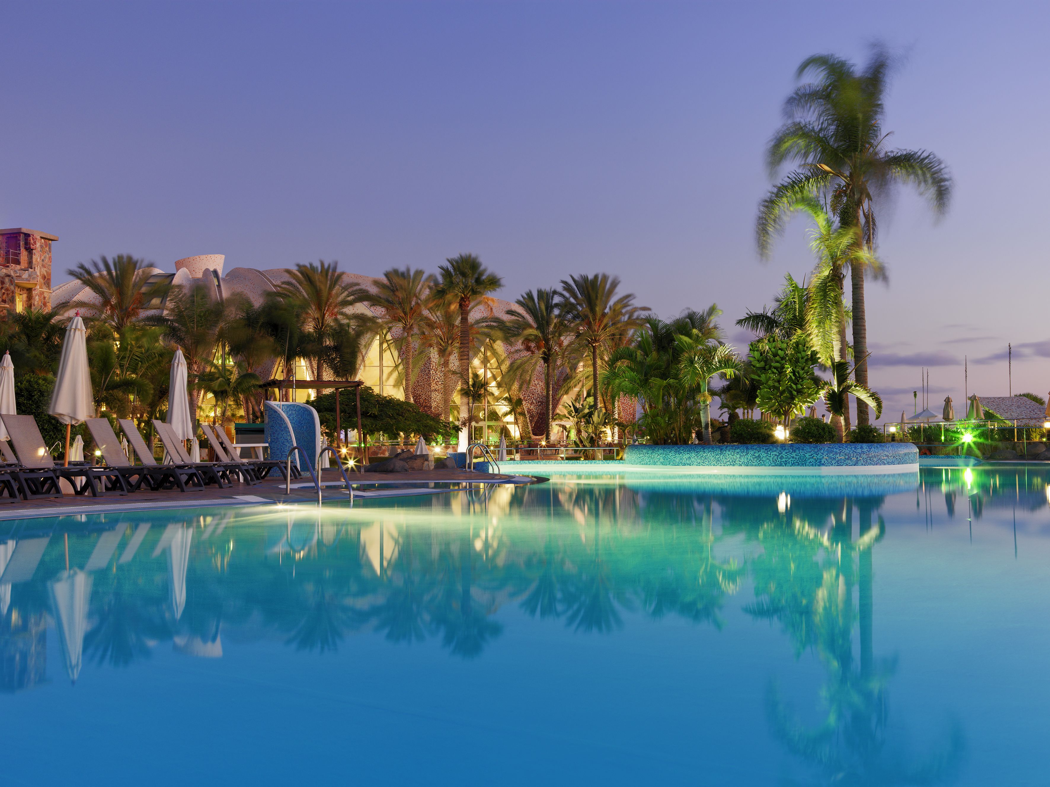 A pool area with lounge chairs and umbrellas, surrounded by palm trees and a building with a dome structure.