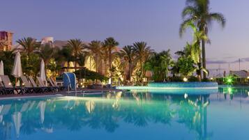 A pool area with lounge chairs and umbrellas, surrounded by palm trees and a building with a dome structure.