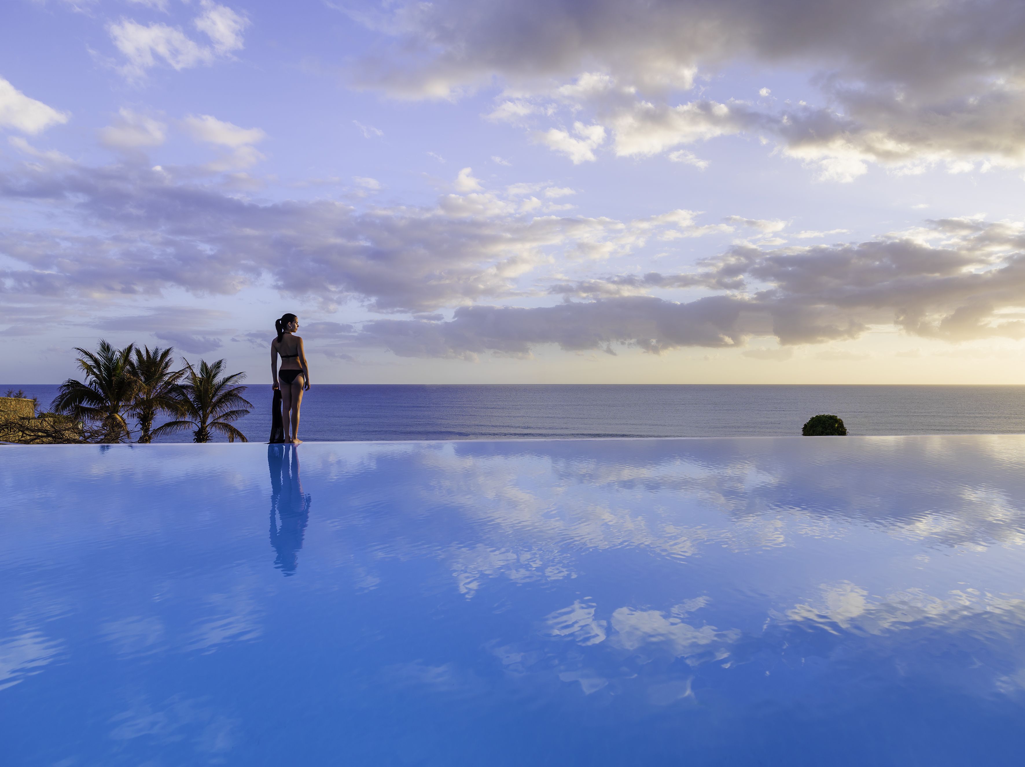 A person standing at the edge of an infinity pool with a view of the ocean and a cloudy sky.