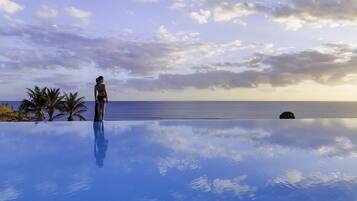 A person standing at the edge of an infinity pool with a view of the ocean and a cloudy sky.