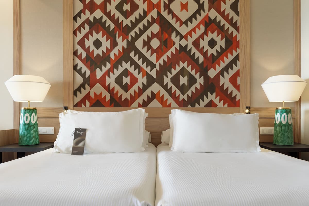 A hotel room with two beds, a patterned wall hanging, and two lamps on nightstands.