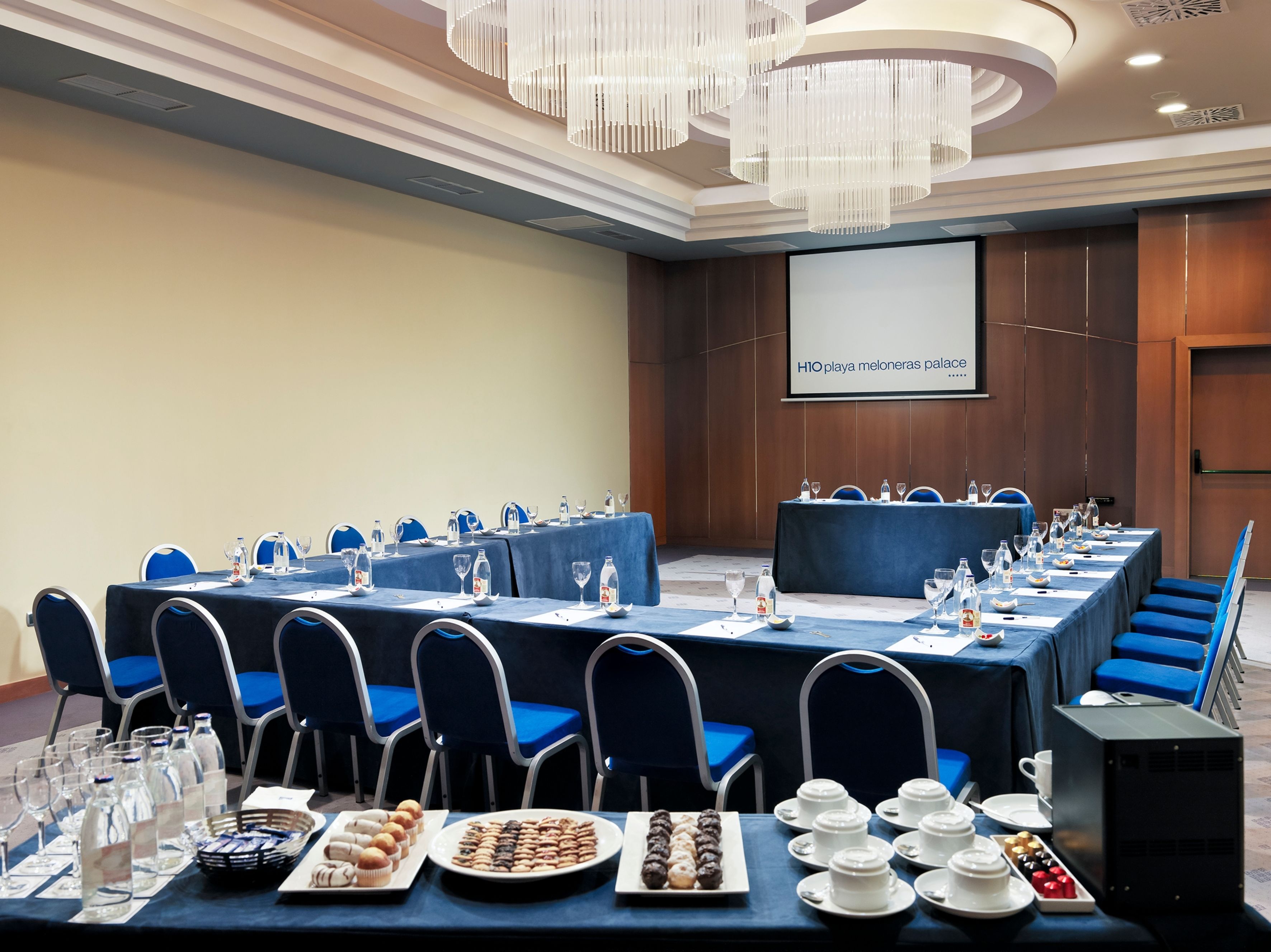 Un salón de conferencias con una larga mesa dispuesta para una reunión, equipado con sillas, botellas de agua y una pantalla de proyector que muestra 