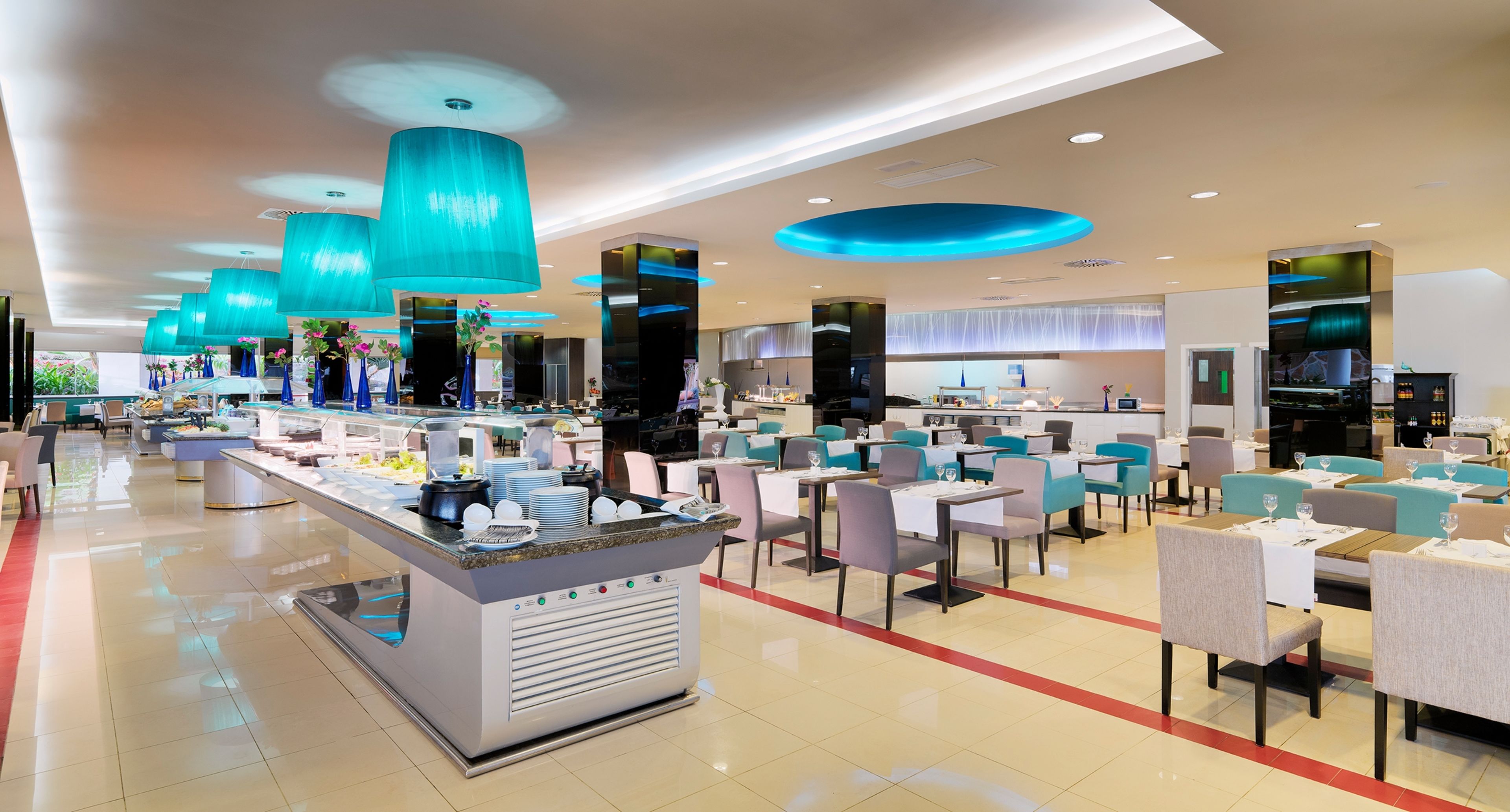 A modern hotel lobby with a central reception desk, multiple dining areas, and a large kitchen in the background.