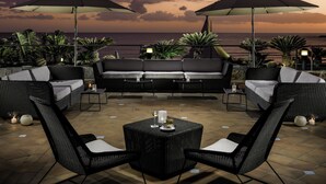 A terrace with a sofa, chairs, and umbrellas overlooking the ocean at sunset.