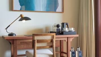 Habitación de hotel con un escritorio de madera, una silla, una mesa redonda con botellas de agua, una cafetera y un cuadro abstracto enmarcado en la pared.