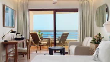 Habitación de hotel con cama, escritorio con lámpara, silla, mesita y vista al océano a través de puertas correderas grandes.
