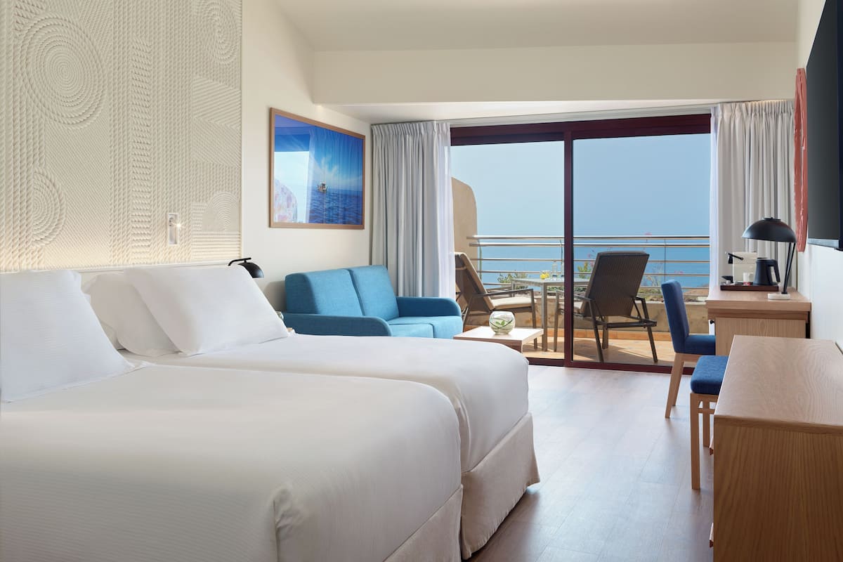 A hotel room with two beds, a sofa, a small table, and a balcony with a view of the sea.