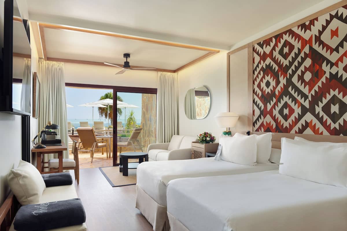 A hotel room with two beds, a desk with a lamp, a TV, a large window with curtains, and a patterned wall.