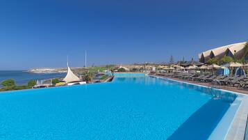A large, clear blue pool with lounge chairs and umbrellas on the right, overlooking the sea.