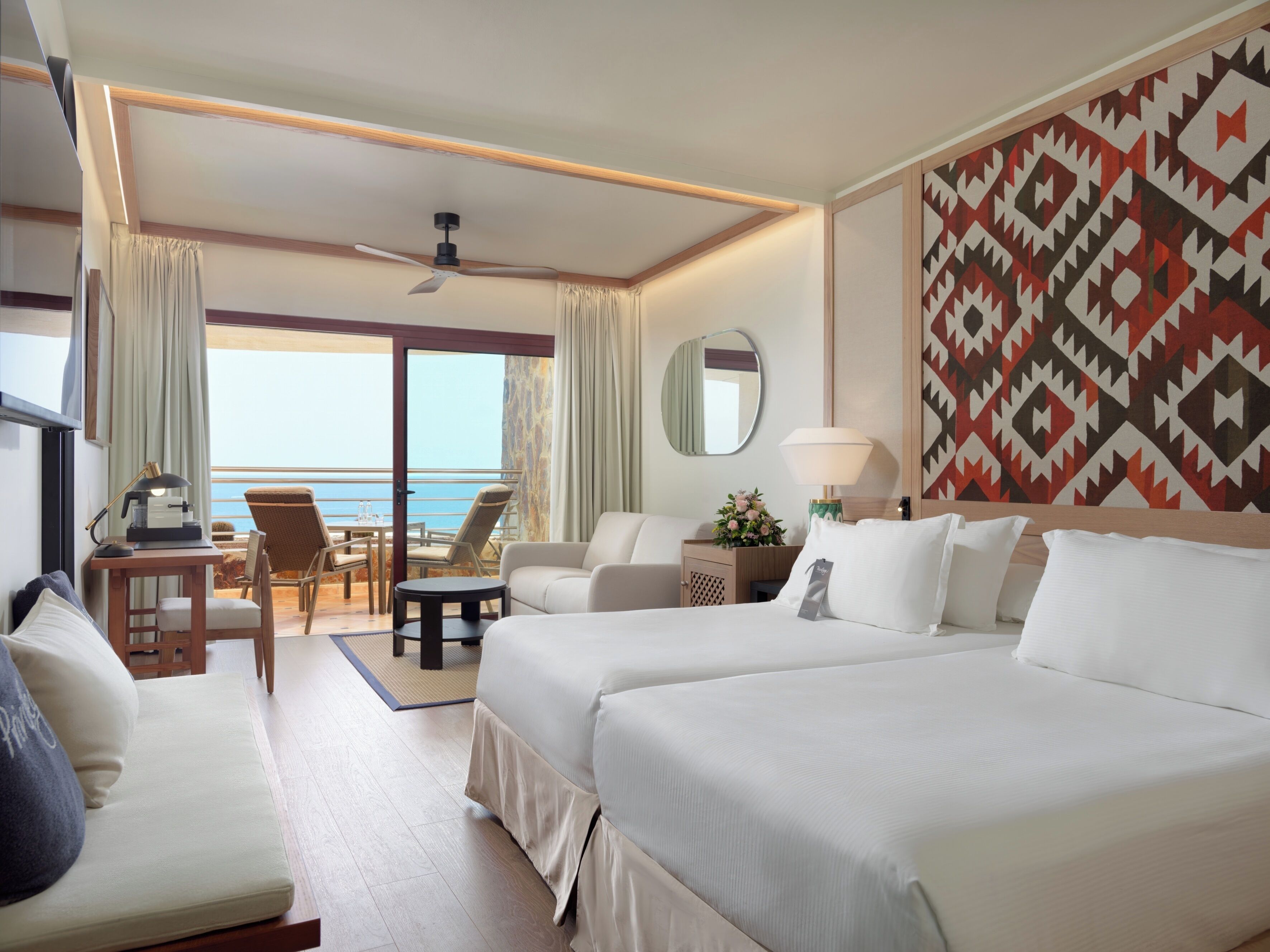 A hotel room with two beds, a desk with a lamp, a chair, a small table, a mirror, and a view of the ocean.