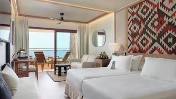 A hotel room with two beds, a desk with a lamp, a chair, a small table, a mirror, and a view of the ocean.
