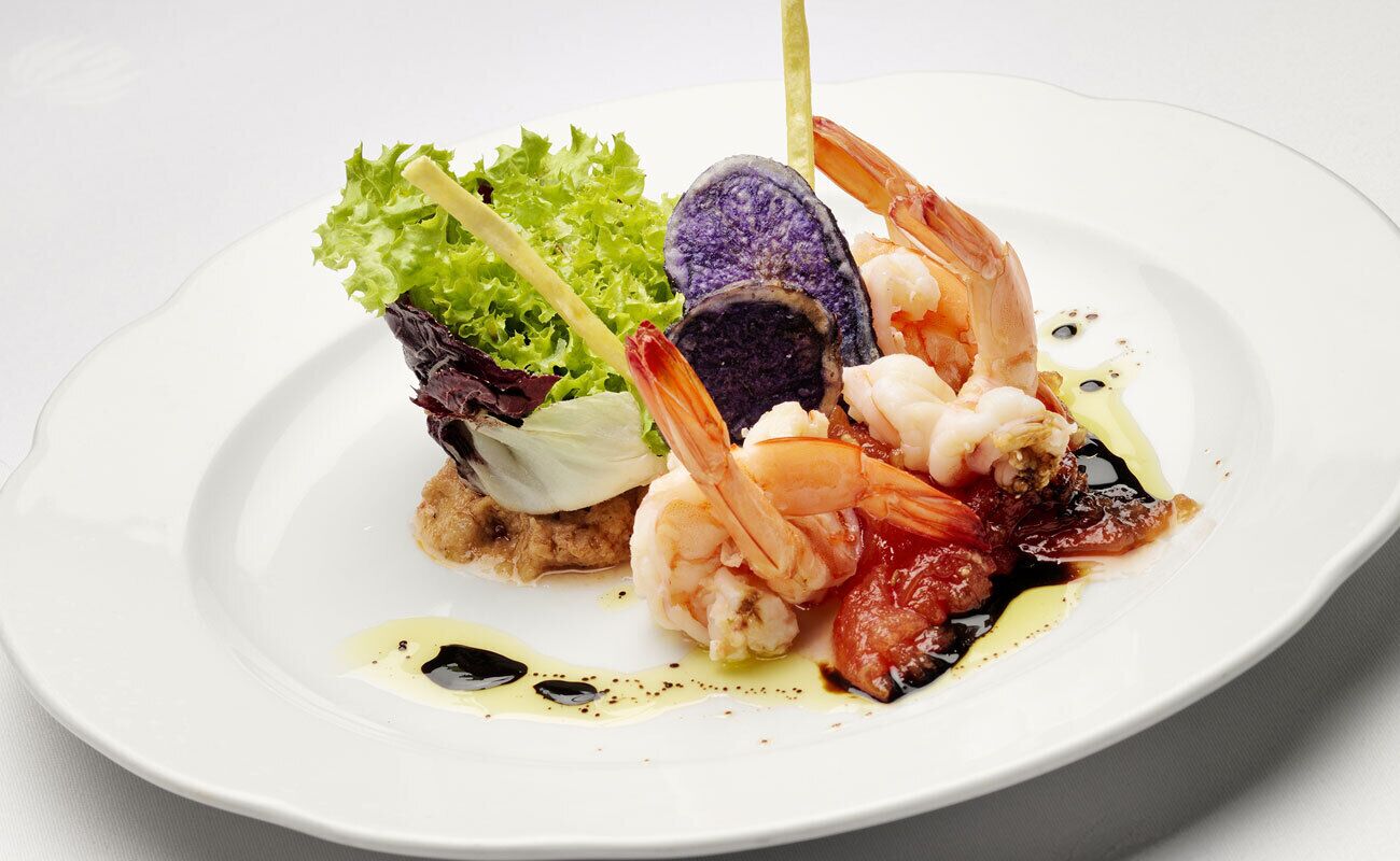 Un plato con mariscos, incluyendo camarones y un elemento morado, posiblemente una verdura o fruta, sobre una cama de hojas verdes.