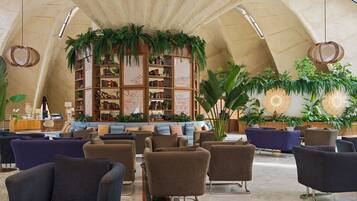 A spacious lobby with a high arched ceiling, a central seating area with sofas and armchairs, and a large bookshelf surrounded by greenery.