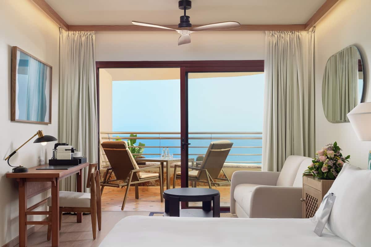 A hotel room with a bed, a desk with a lamp, a chair, a small table, and a view of the ocean.