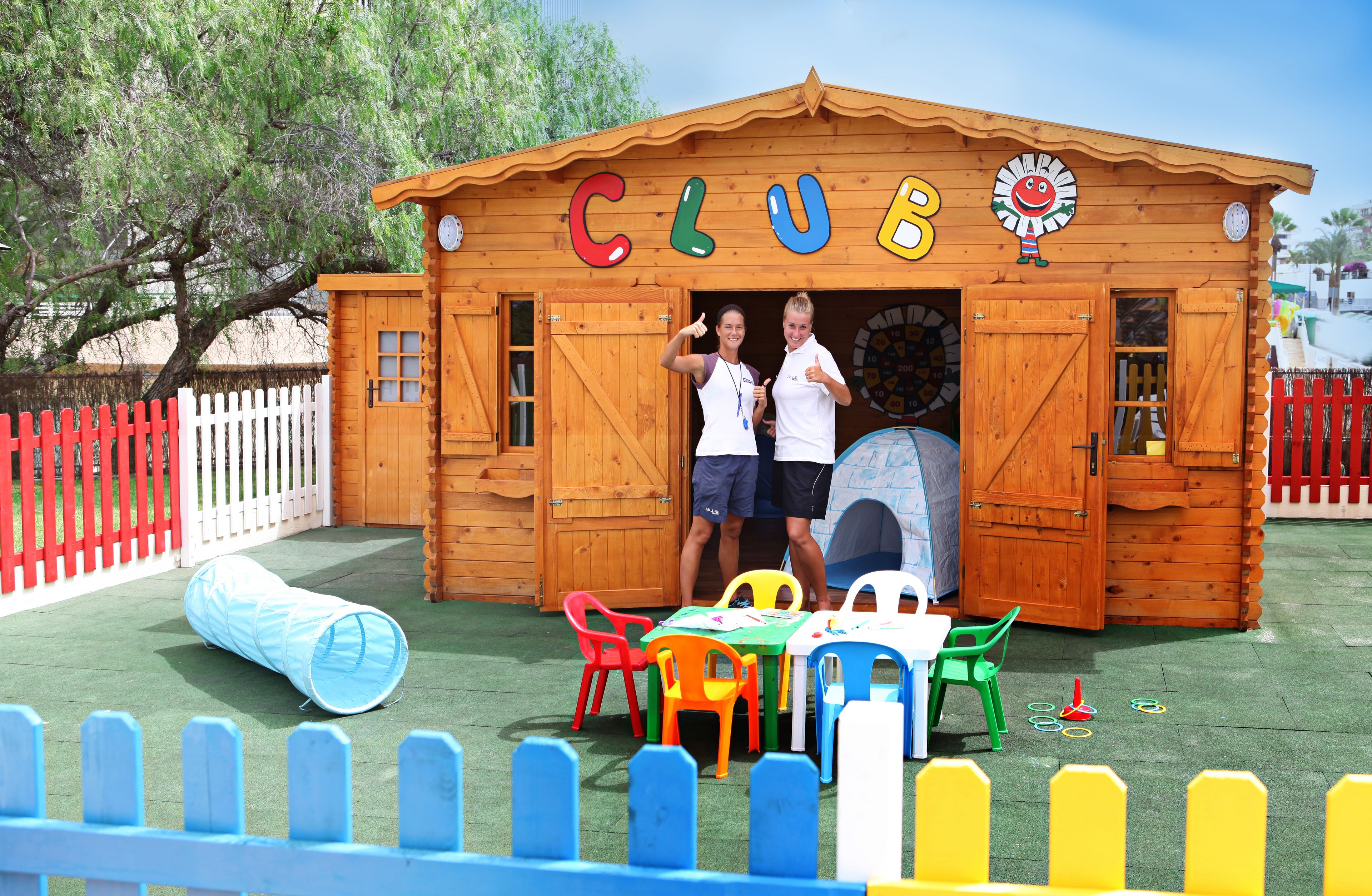 A wooden clubhouse with the word "CLUB" on the front, a red and white picket fence, and colorful plastic chairs and tables outside.