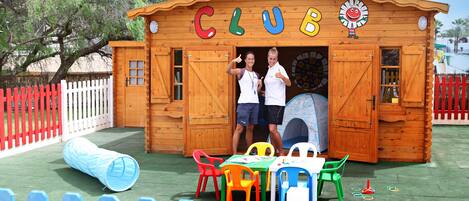 A wooden clubhouse with the word "CLUB" on the front, a red and white picket fence, and colorful plastic chairs and tables outside.
