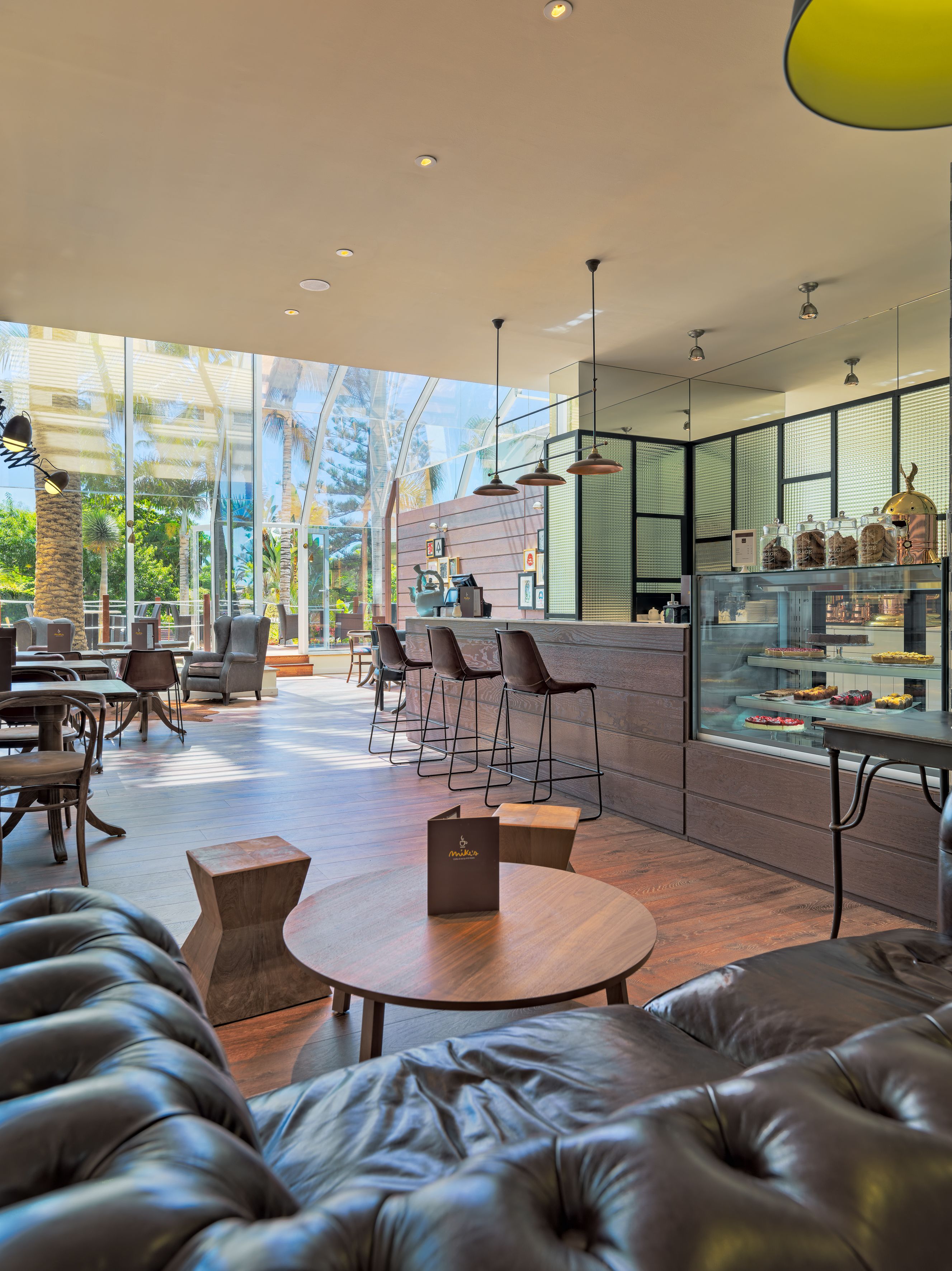 Een modern café met lederen zitplaatsen, een glazen vitrine met gebak en uitzicht op palmbomen door grote ramen.