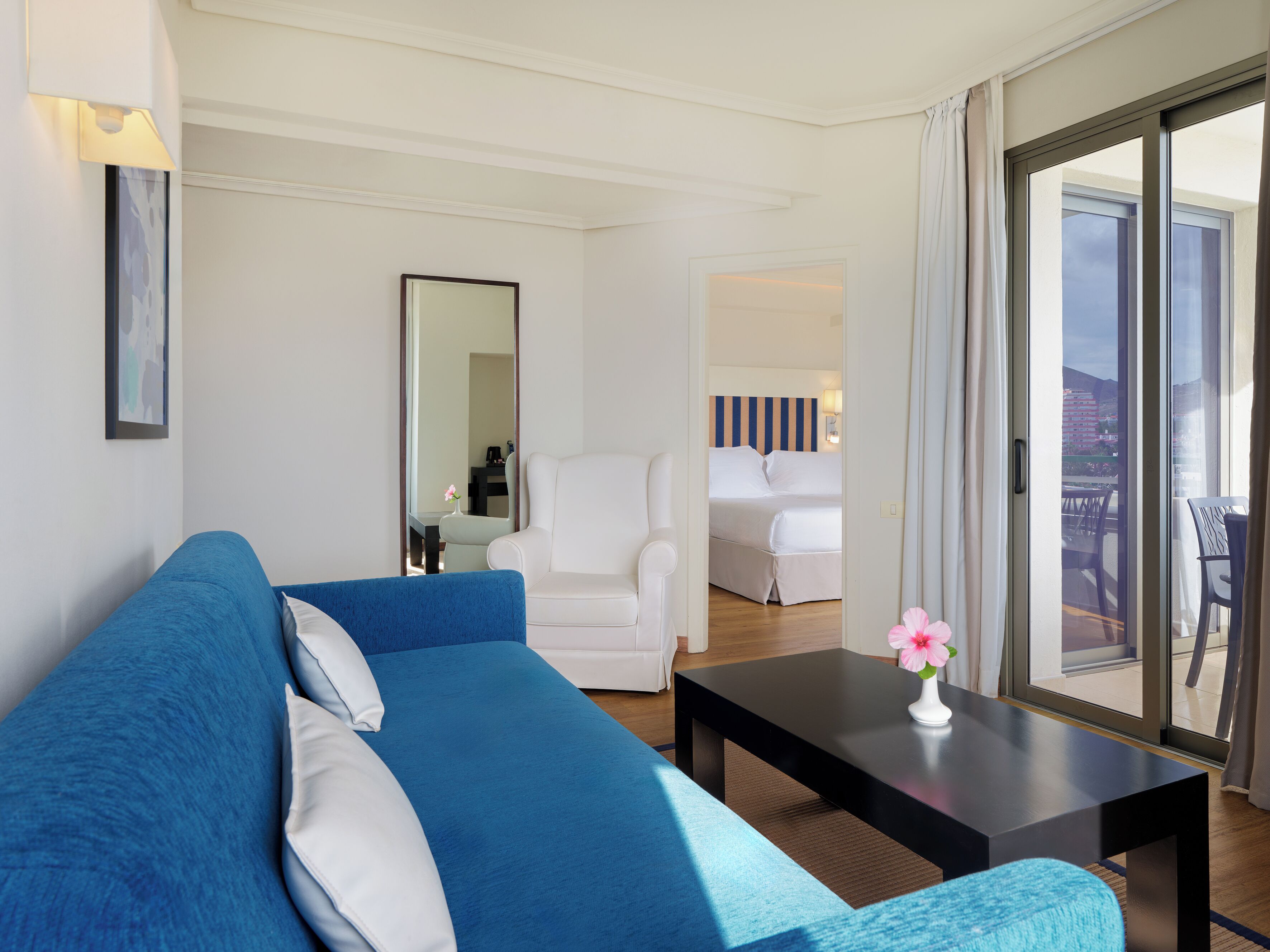 A modern hotel room with a blue sofa, a white armchair, a small table with a vase, and a large sliding glass door leading to a balcony.