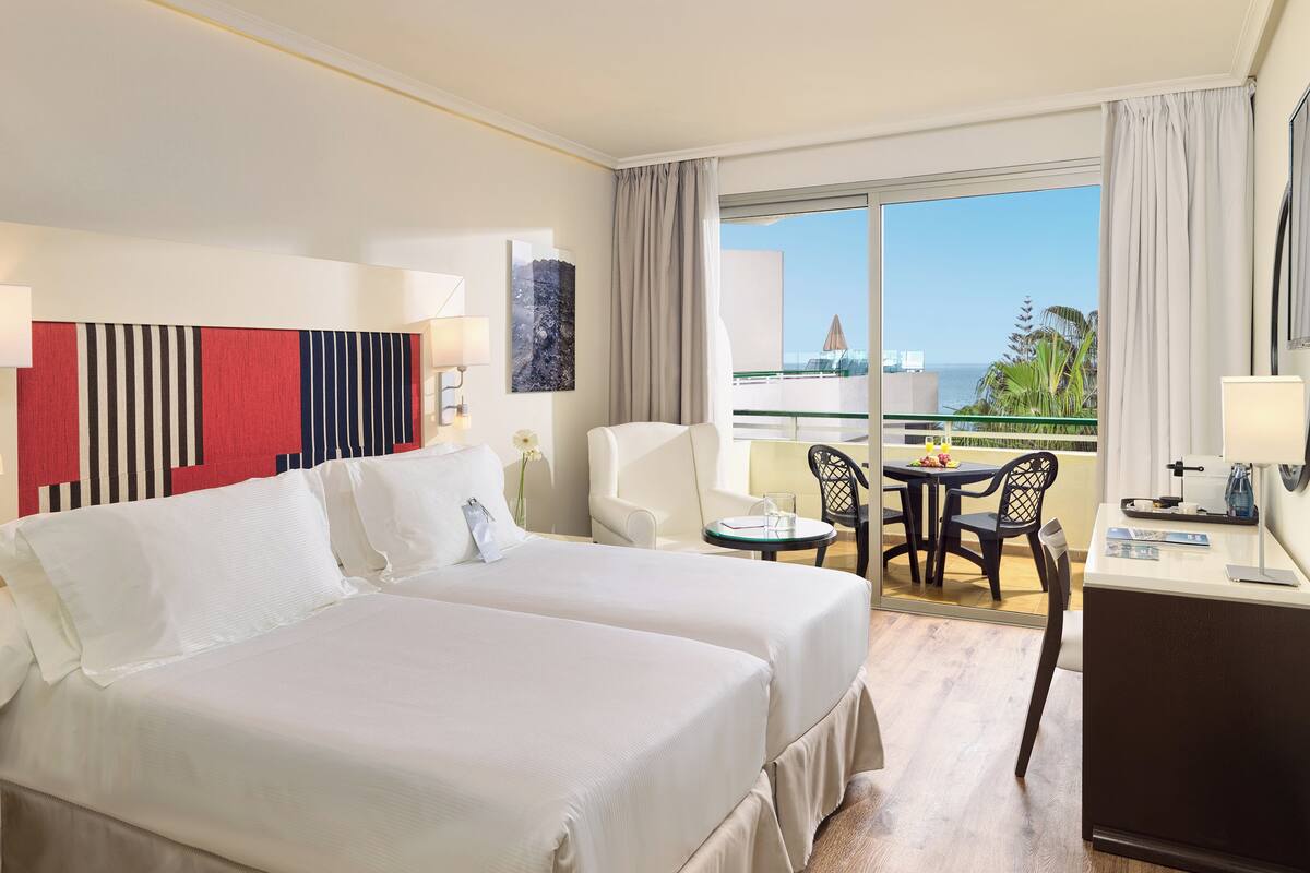 A hotel room with two beds, a desk, a chair, a small table, and a balcony with a view of the ocean.