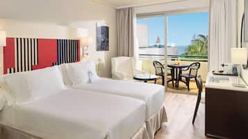 Habitación de hotel con dos camas, un escritorio, una silla, una mesita y un balcón con vistas al océano.