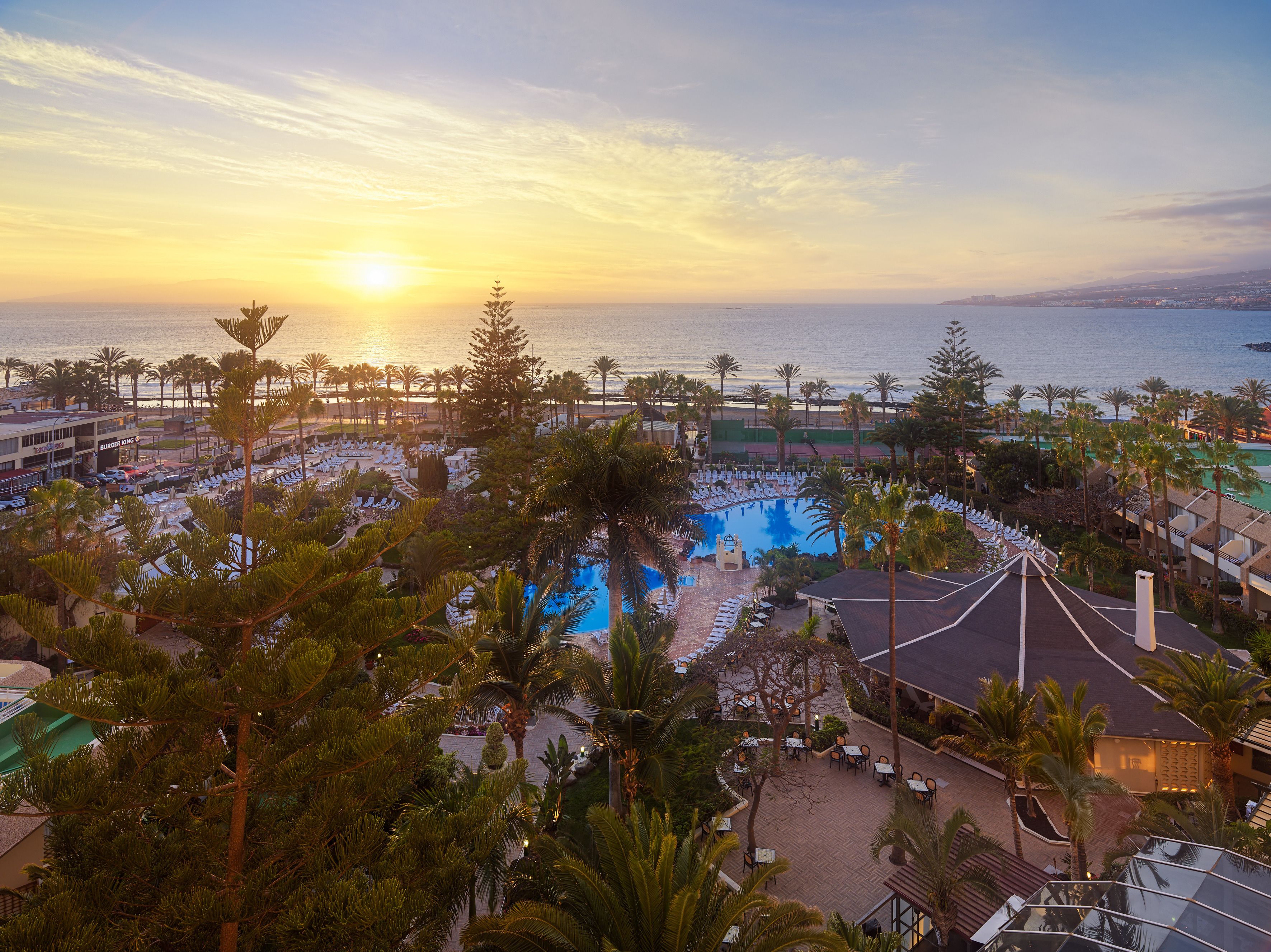 A resort with a pool, palm trees, and a view of the ocean at sunset.