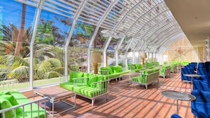 Un espacio interior amplio con techo de vidrio, asientos verdes y azules, y piso de madera.