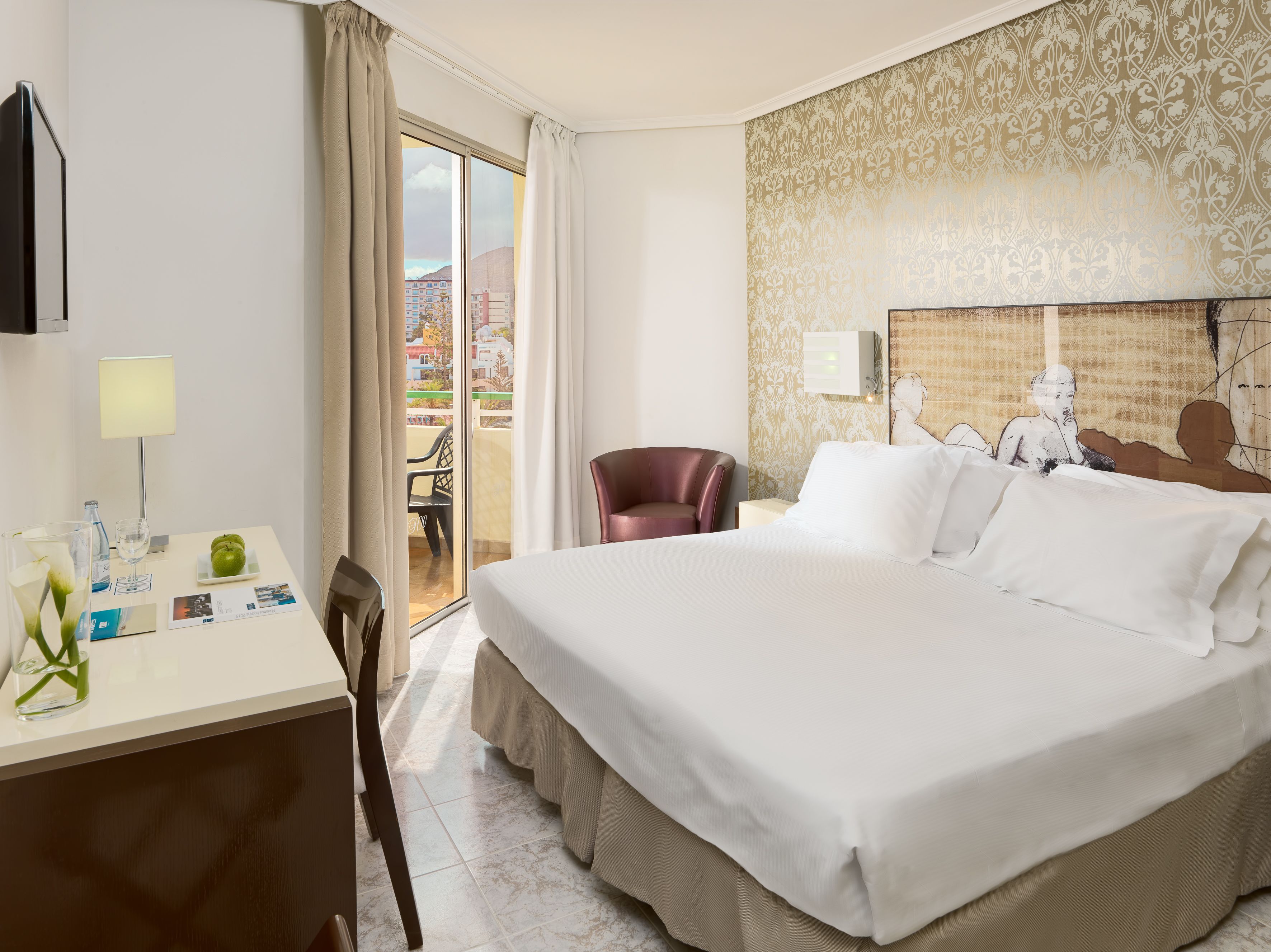 Habitación de hotel con una cama grande, un escritorio con lámpara, una silla y vistas a edificios a través de una puerta corredera.
