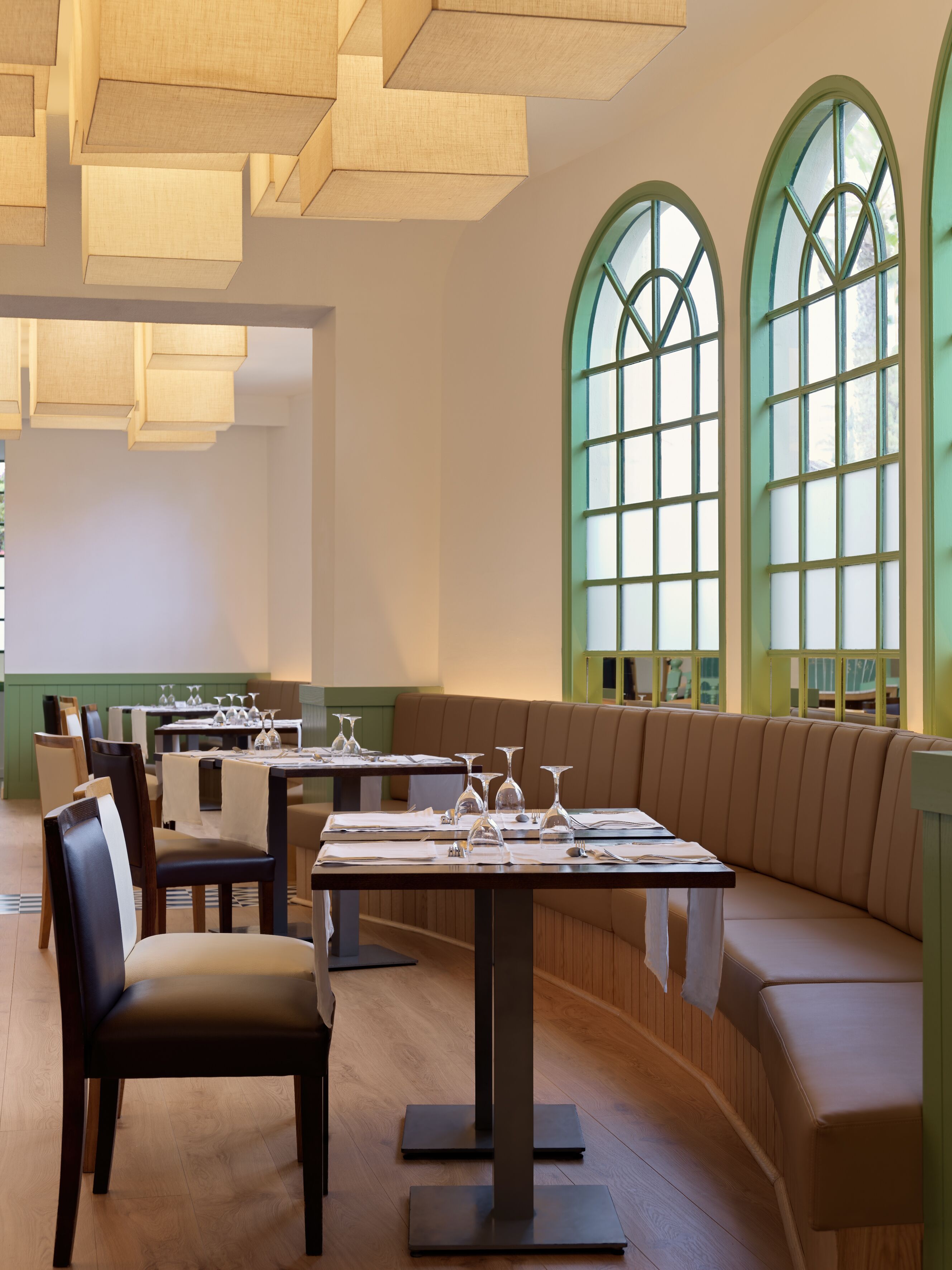 A restaurant with a modern interior, featuring a long dining table with chairs, a cushioned bench, and large arched windows.