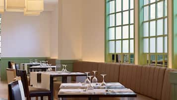 A restaurant with a modern interior, featuring a long dining table with chairs, a cushioned bench, and large arched windows.