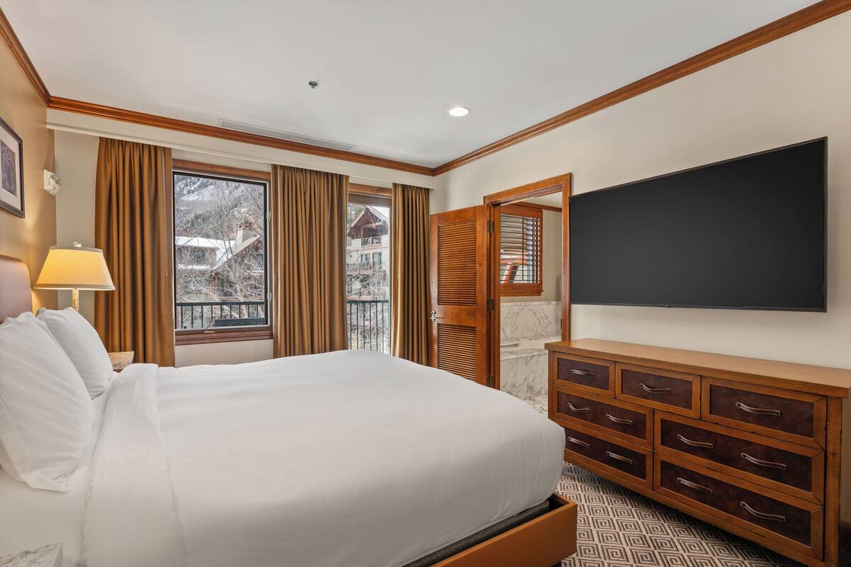 A hotel room with a large bed, a flat-screen TV, a wooden dresser, and a window with curtains.