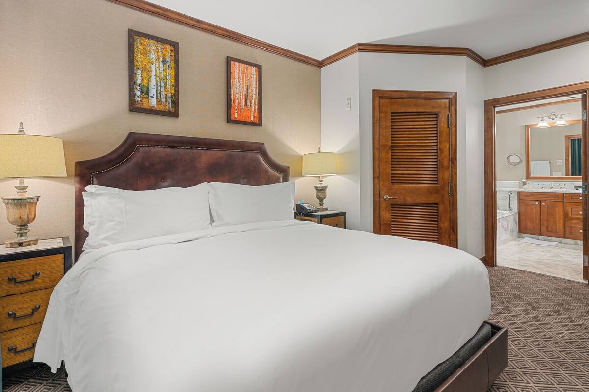 A hotel room with a large bed, two bedside lamps, a wooden headboard, a nightstand, a dresser, and a bathroom visible through an open door.