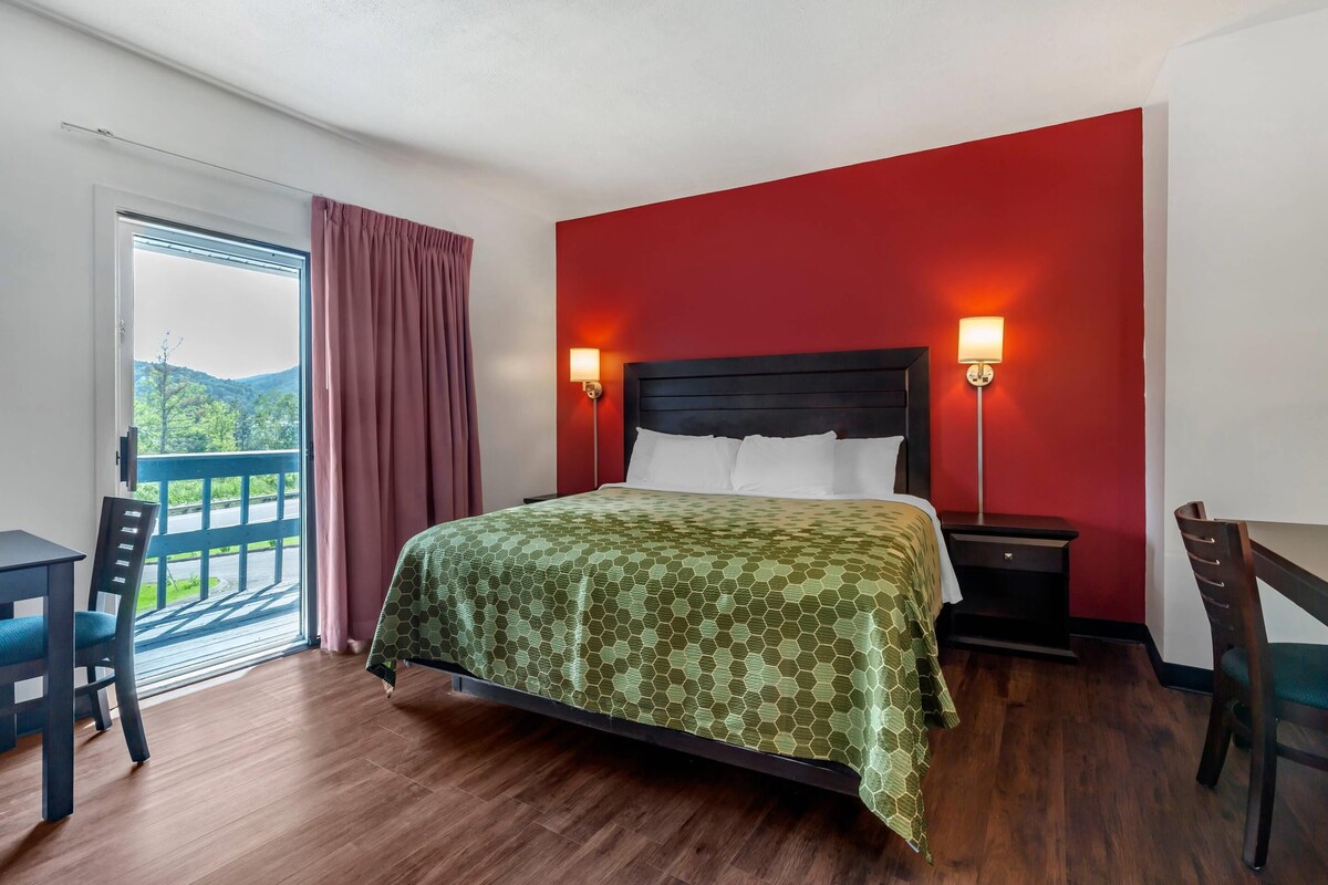 A hotel room with a bed, a desk, and a chair. There is a window with a view of greenery outside.
