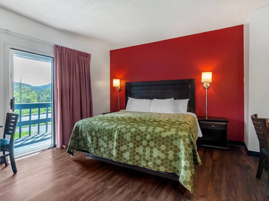 Ein Hotelzimmer mit einem Bett, einem Schreibtisch und einem Stuhl. Ein Fenster bietet Blick auf eine grüne Umgebung.