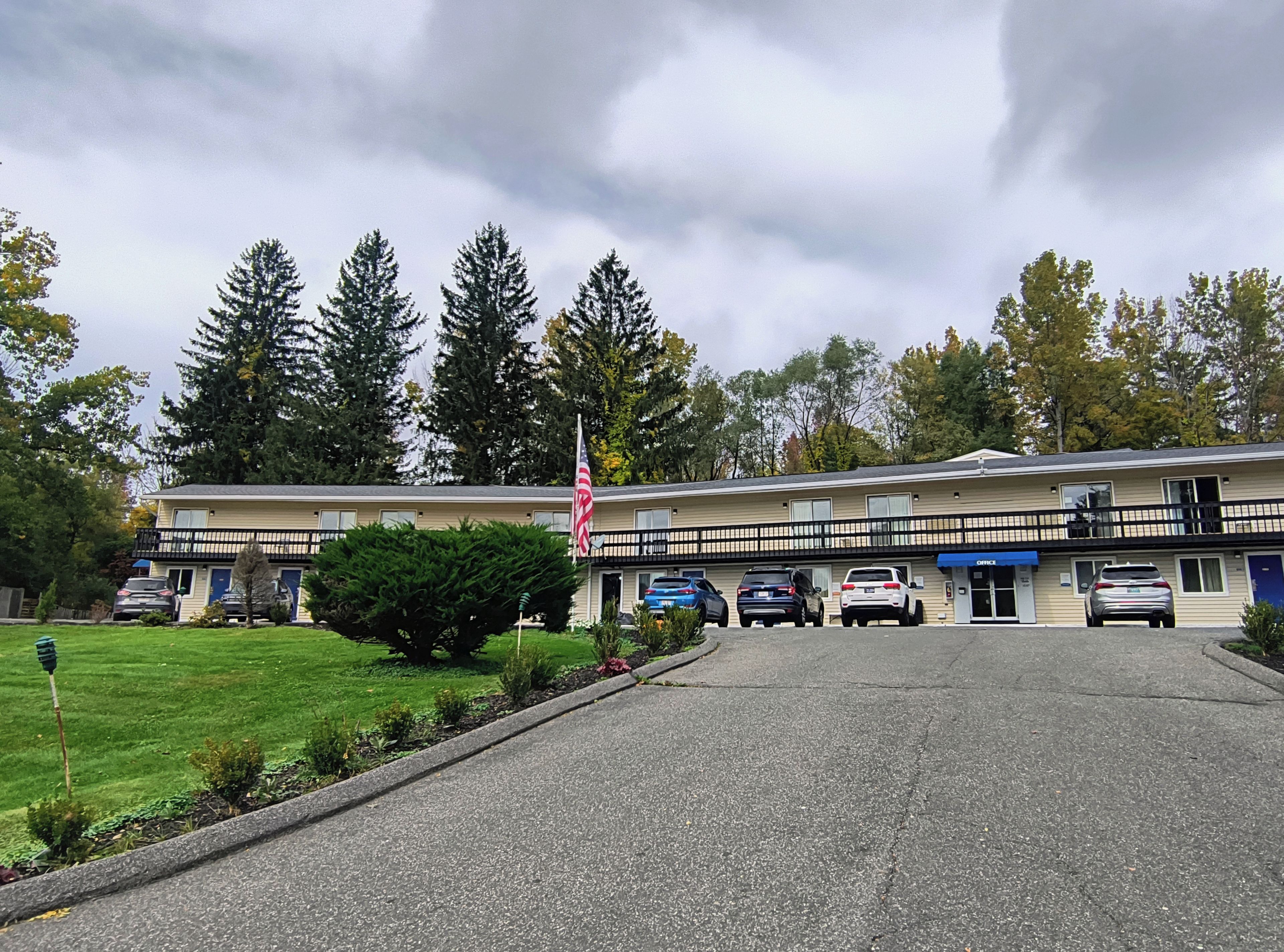 Een motel met een centrale ingang, een vlag en een parkeerplaats met meerdere auto's.
