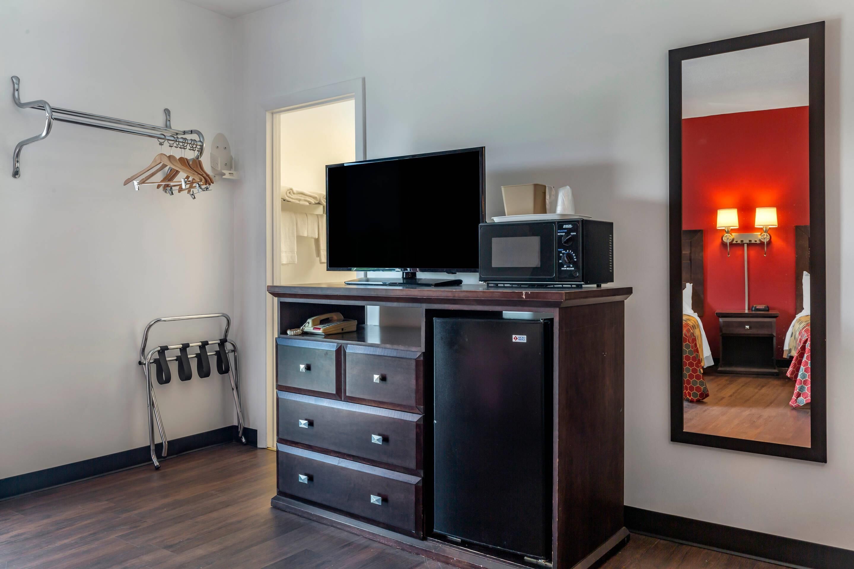 A hotel room with a flat-screen TV, a microwave, a mini-fridge, a wooden dresser, a coat rack, and a mirror reflecting a red-lit room.