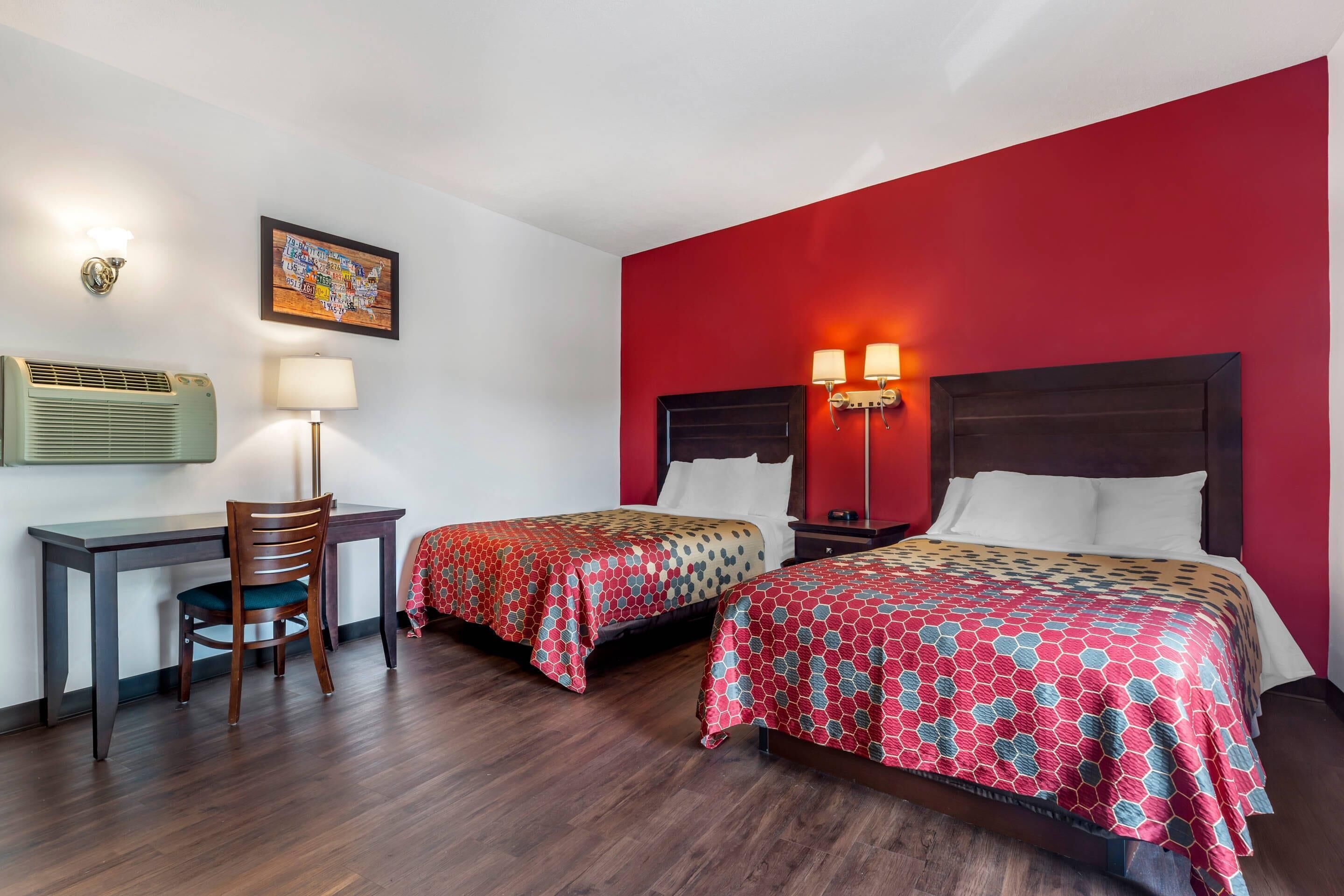 A hotel room with two beds, a desk, a chair, and a wall-mounted air conditioner.