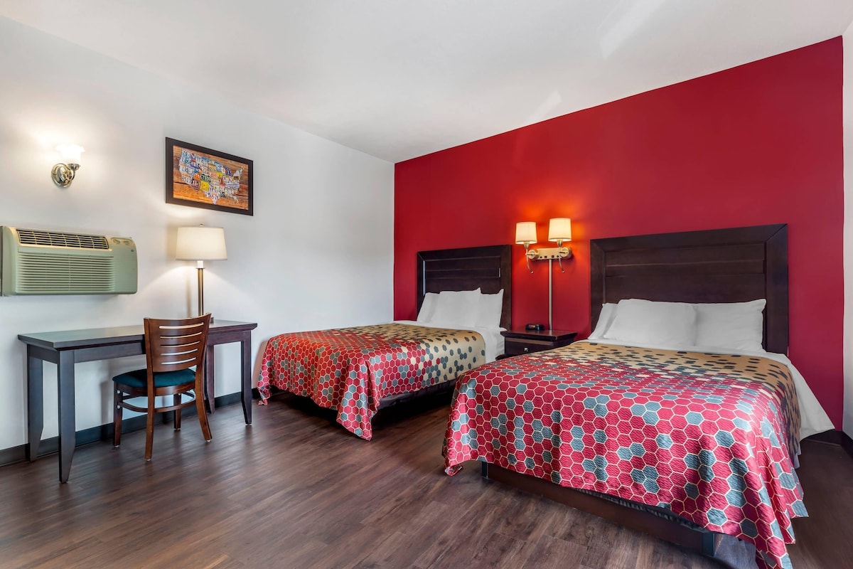 A hotel room with two beds, a desk, a chair, and a wall-mounted air conditioner.