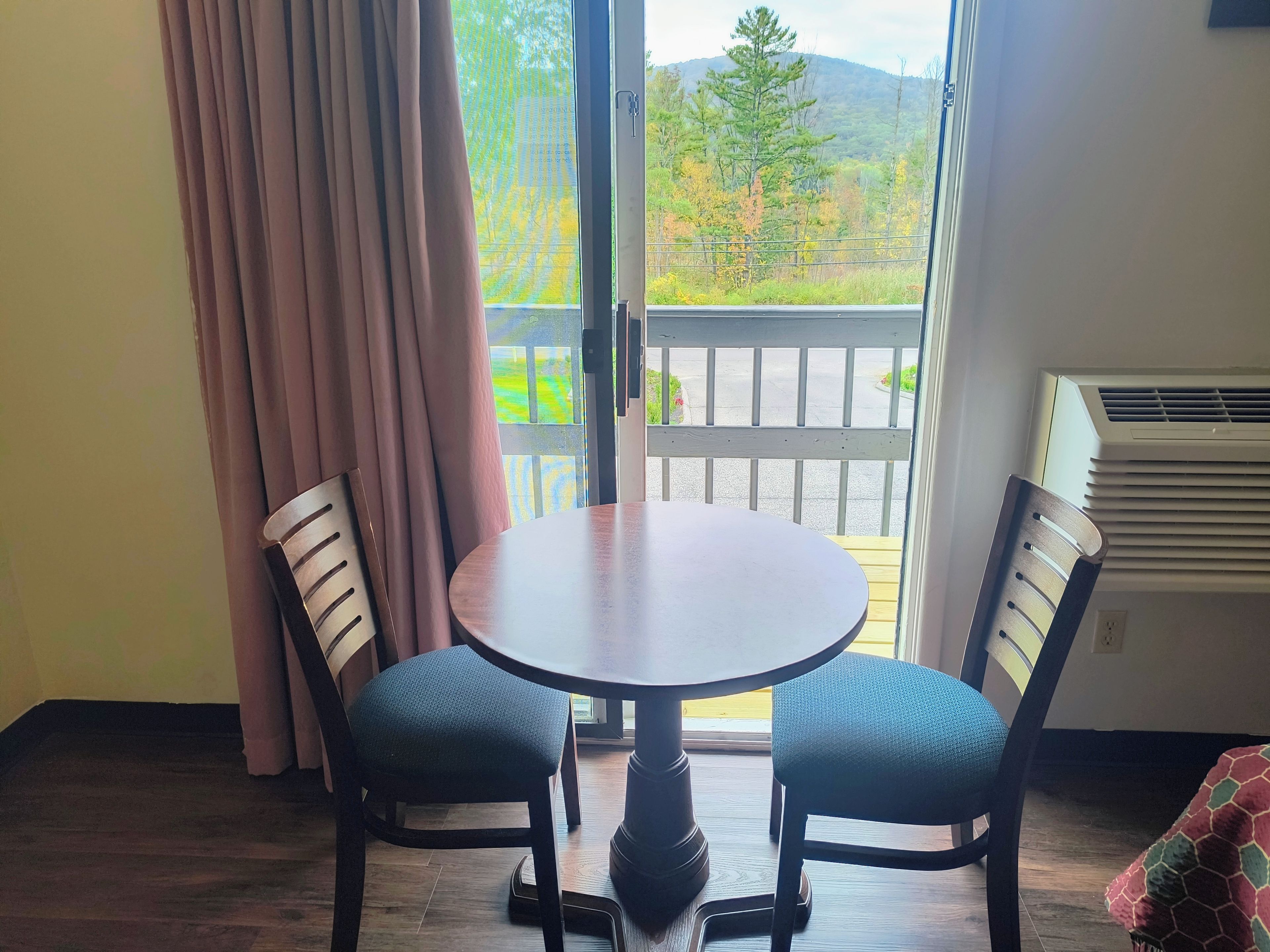 A room with a round table and two chairs, a window with a view of trees and a building, and an air conditioning unit.
